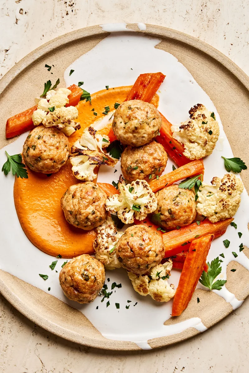 Romesco sauce on a plate piled with Chicken meatballs and roasted carrots and cauliflower that were all baked on a sheet pan