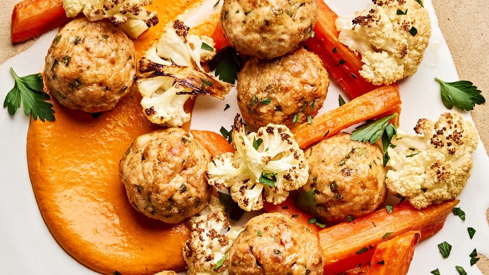 Romesco sauce on a plate piled with Chicken meatballs and roasted carrots and cauliflower that were all baked on a sheet pan