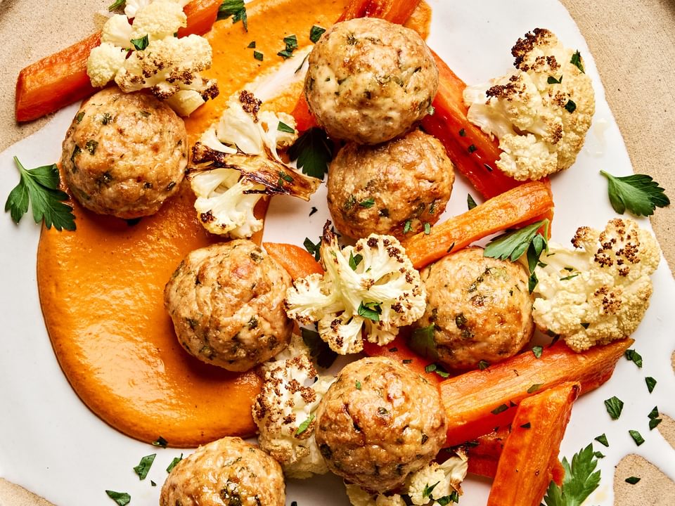 Romesco sauce on a plate piled with Chicken meatballs and roasted carrots and cauliflower that were all baked on a sheet pan