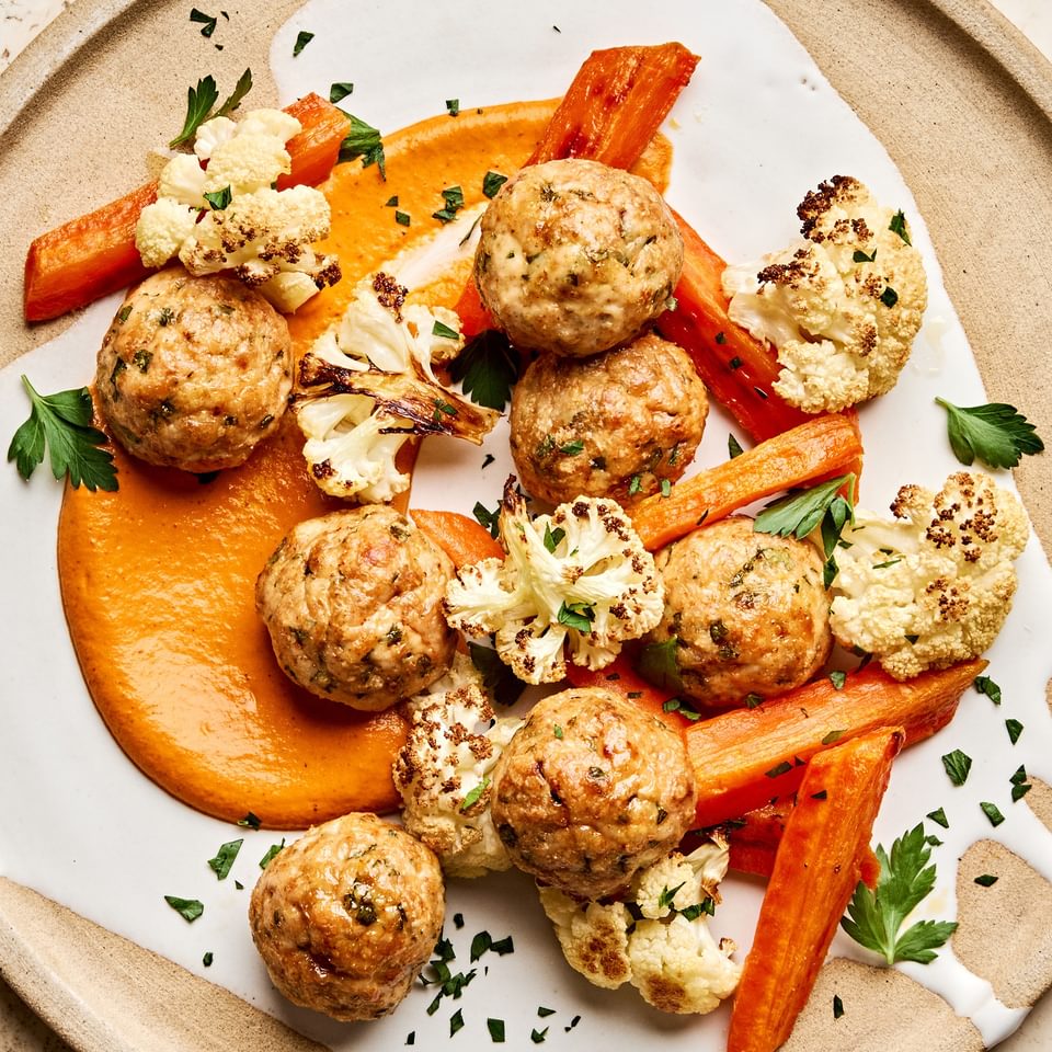 Romesco sauce on a plate piled with Chicken meatballs and roasted carrots and cauliflower that were all baked on a sheet pan