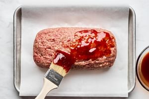 A perfectly formed meatloaf on a parchment-lined baking sheet, being brushed with a savory, sweet sauce for added flavor.