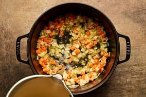 Chicken broth being poured into a pot with onion, garlic and carrot in a butter and flour roux.