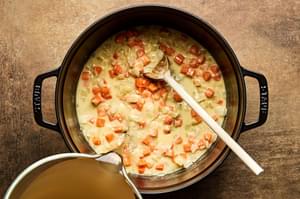 Chicken broth being stirred into a pot with onion, garlic and carrot in a butter and flour roux.
