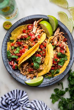 Three chicken tacos shown in a metal pie pan with salsa and limes next to a linen and fresh cilantro