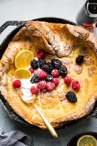 Homemade Dutch baby pancake in a cast iron skillet topped with whipped cream, powdered sugar, berries and lemon