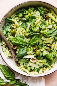 Green goddess pasta salad with basil, green olives, asparagus, snap peas and cucumbers in a a bowl with a wooden spoon