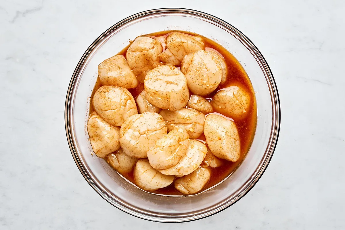 Raw sea scallops marinating in savory sauce in a glass bowl before grilling on a grill.