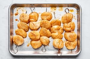 Marinated scallops threaded on metal skewers ready for grilling on a baking sheet.