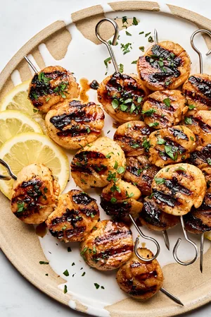 Perfectly grilled scallops on metal skewers with lemon slices and fresh herbs on a plate.