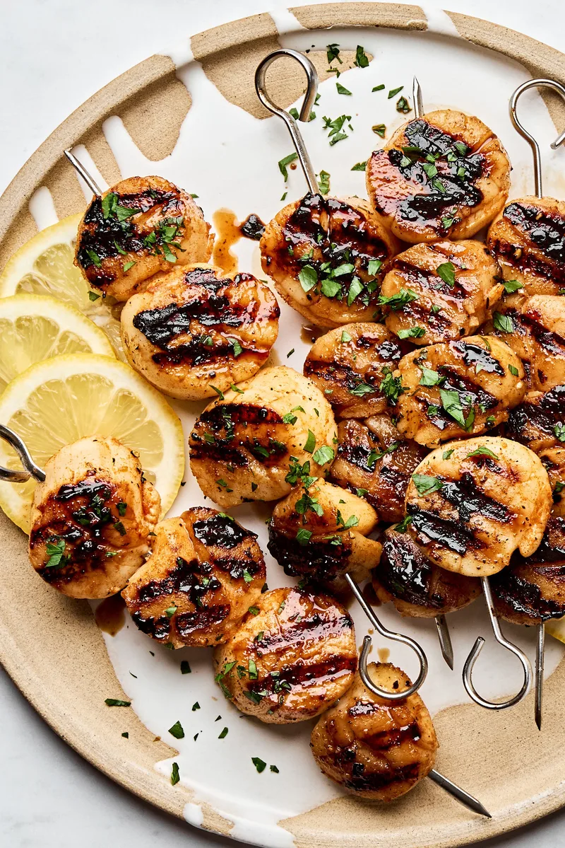 Perfectly grilled scallops on metal skewers with lemon slices and fresh herbs on a plate.