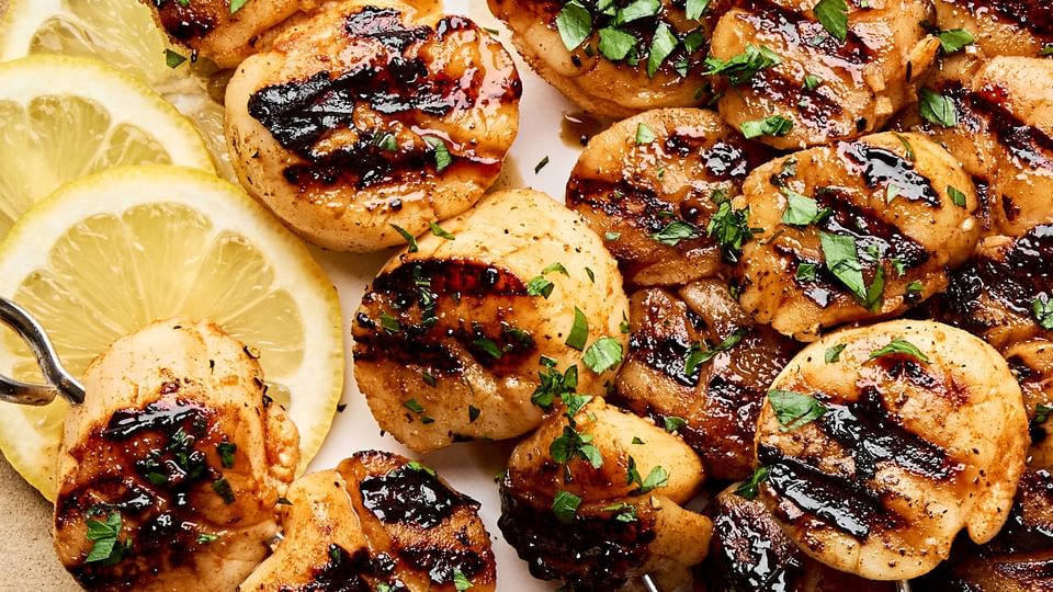 Perfectly grilled scallops on metal skewers with lemon slices and fresh herbs on a plate.