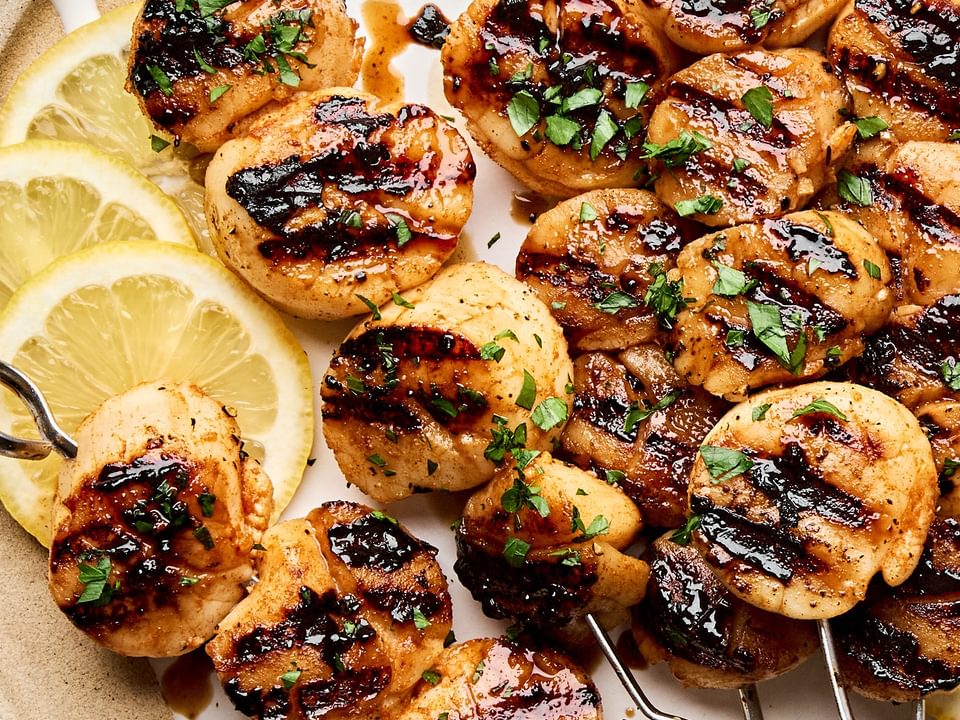 Perfectly grilled scallops on metal skewers with lemon slices and fresh herbs on a plate.