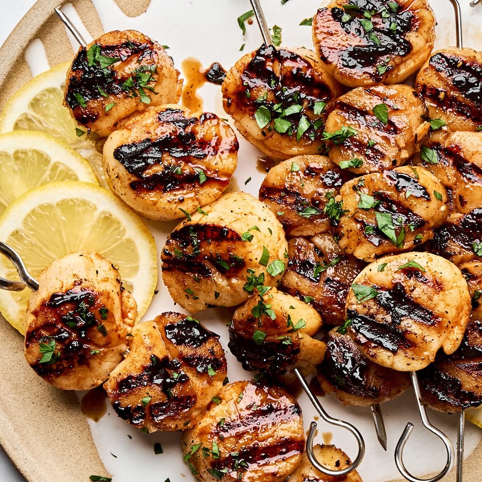 Perfectly grilled scallops on metal skewers with lemon slices and fresh herbs on a plate.