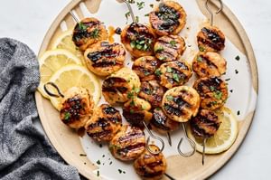 Perfectly grilled scallops on metal skewers with lemon slices and fresh herbs on a plate.