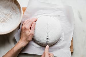 hands scoring sourdough bread dough using a bread lame