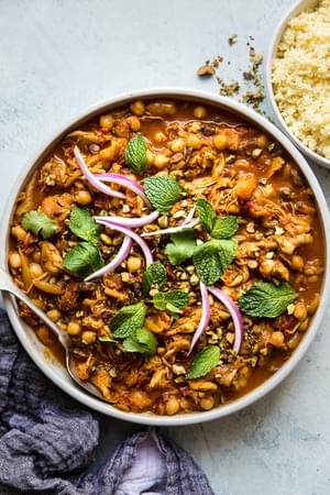 chicken tagine with a bowl of cous cous next to it topped with fresh herbs and purple onions