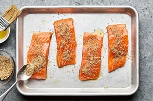 4 raw salmon fillets on a baking sheet being rubbed with oil and sprinkled with seasoning, ready to be cooked.
