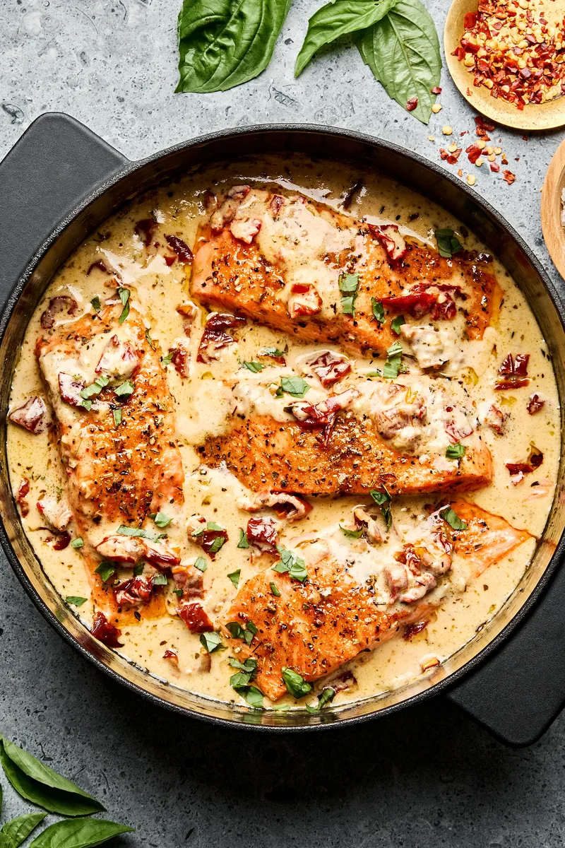 Marry Me Salmon fillets simmering in a creamy, sun-dried tomato sauce inside a skillet, garnished with basil and Parmesan.