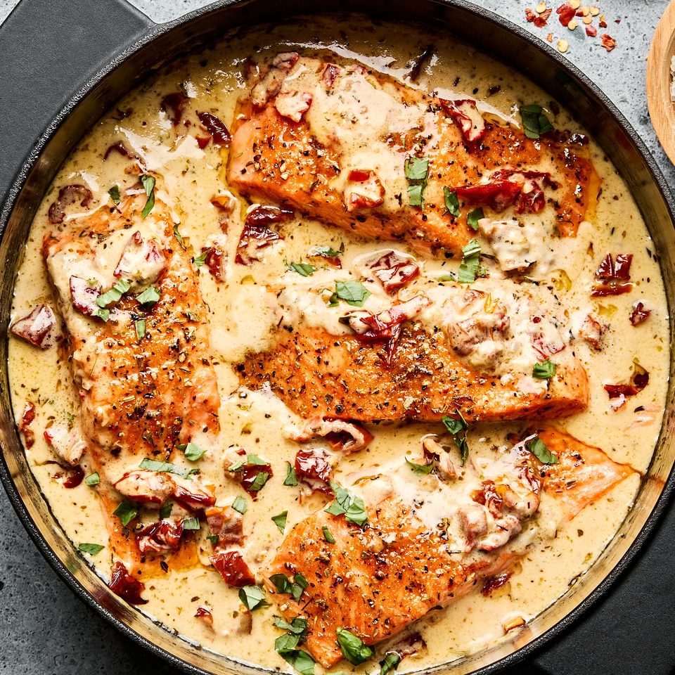 Marry Me Salmon fillets simmering in a creamy, sun-dried tomato sauce inside a skillet, garnished with basil and Parmesan.