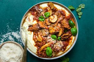 a bowl of mushroom ragù served over creamy polenta topped with basil leaves next to a bowl of grated parmesan