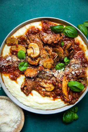 a bowl of mushroom ragù served over creamy polenta topped with basil leaves next to a bowl of grated parmesan
