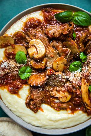 a bowl of mushroom ragù served over creamy polenta topped with basil leaves next to a bowl of grated parmesan