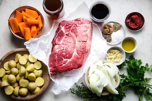 Ingredients for pot roast including potatoes, chuck roast, carrots, onions, tomato paste, red wine and broth.