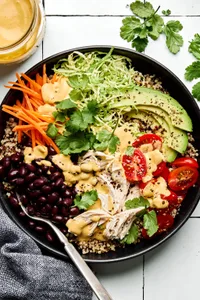 a quinoa bowl with chicken, black beans, cilantro, carrots, tomatoes, avocado, brussel sprouts drizzled with special sauce
