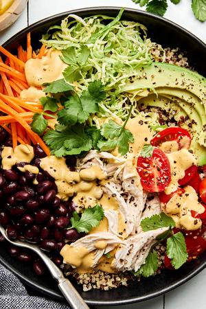 a quinoa bowl with chicken, black beans, cilantro, carrots, tomatoes, avocado, brussel sprouts drizzled with special sauce