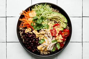 a quinoa bowl with chicken, black beans, cilantro, carrots, tomatoes, avocado, brussel sprouts drizzled with special sauce