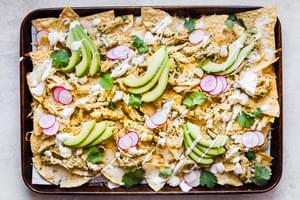 salsa verde nachos topped with sour cream, cilantro, avocado and radishes shown on a baking sheet