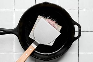 a smash burger being smashed flat with parchment paper and a metal spatula in a cast iron skillet