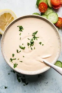 homemade tahini sauce in a bowl with a spoon next to tomatoes, cucumbers and lemons