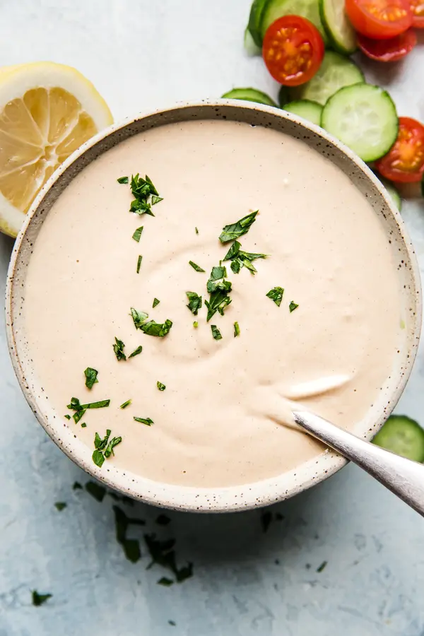 homemade tahini sauce in a bowl with a spoon next to tomatoes, cucumbers and lemons