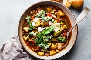 a bowl of homemade vegetable soup topped with fresh basil and parmesan cheese next to fresh bread