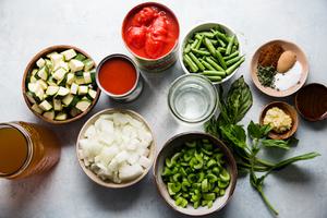 ingredients for vegetable soup in bowls including whole canned tomatoes, onions, celery, spices, green beans, basil & spices.