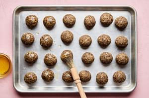 Unbaked vegetarian meatballs on a baking sheet, being brushed with olive oil using a pastry brush, ready to go into the oven.