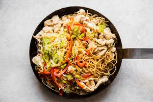 chicken, chow mein noodles and stir fried veggies being tossed in sauce in a skillet