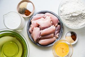 Chicken thighs in a metal dish with oil, eggs, spices along side a pitcher of buttermilk and a bowl of flour.