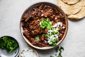 Beef Birria in a bowl topped with chopped onions and cilantro surrounded by tortillas and bowls of extra cilantro and onions