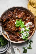 a bowl of homemade shredded beef birria topped with chopped onion and cilantro next to tortillas for serving