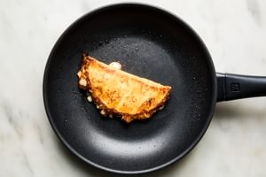 A birria taco being fried in a skillet, with crispy edges forming on the tortilla and rich, red consommé seeping out