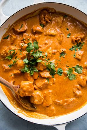 a pot of butter chicken topped with fresh cilantro being stirred with a wooden spoon