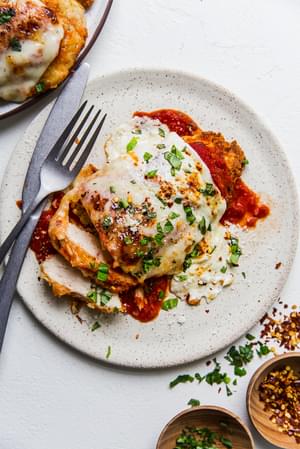 homemade chicken parmesan on a plate with marinara, basil and fresh mozzarella with a fork and knife