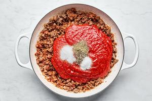 crushed tomatoes and spices being added to cooked ground beef, chopped onions, minced garlic and tomato paste in a skillet