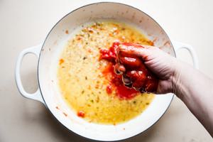 hand crushing canned whole tomatoes over a braising dish with butter garlic, anchovies and white wine