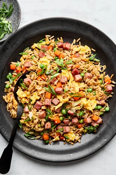 A bowl of ham fried rice, featuring fluffy rice mixed with diced ham, scrambled egg, peas & carrots, topped with green onions