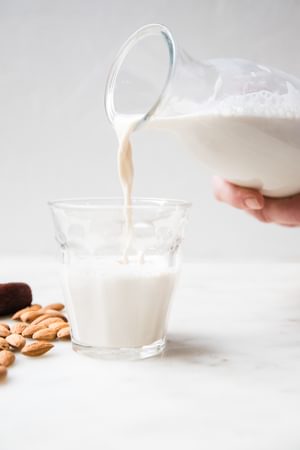 a carafe of homemade almond milk being poured into a glass surrounded by almonds and dates on the counter