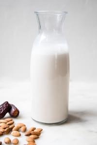 Homemade almond milk in a carafe on the counter surrounded by almonds and dates