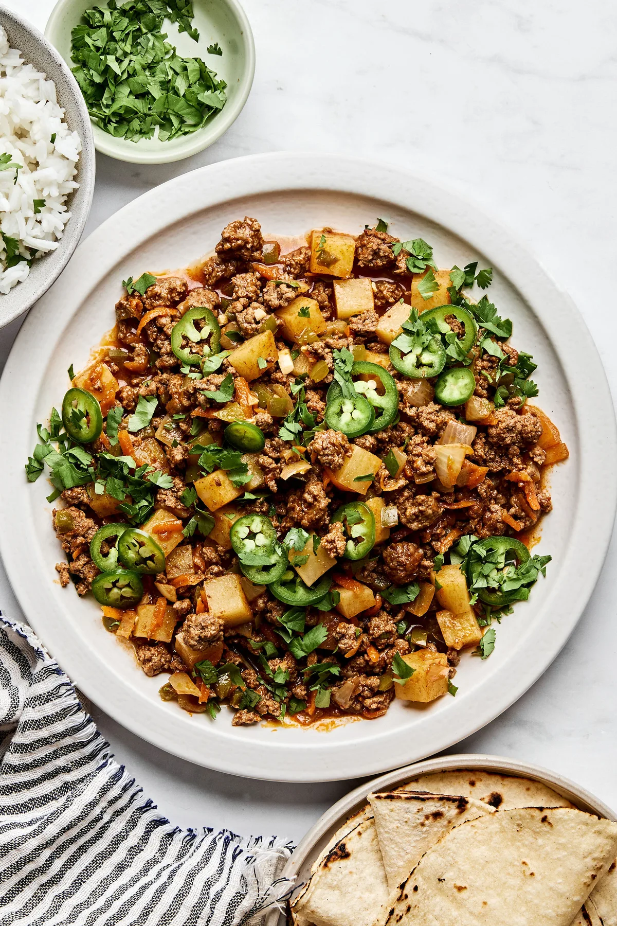 A bowl of picadillo topped with fresh cilantro and jalapeños, served with rice and a warm tortillas on the side